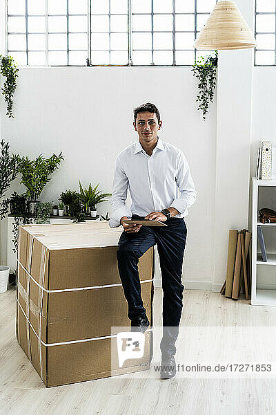 Selbstbewusster Geschäftsmann  der ein Klemmbrett hält und auf einem Karton im Büro sitzt