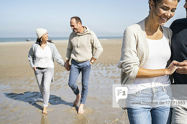 Lächelnde Frau  die einen Mann an der Hand hält  während sie mit einem Paar im Hintergrund am Strand spazieren geht