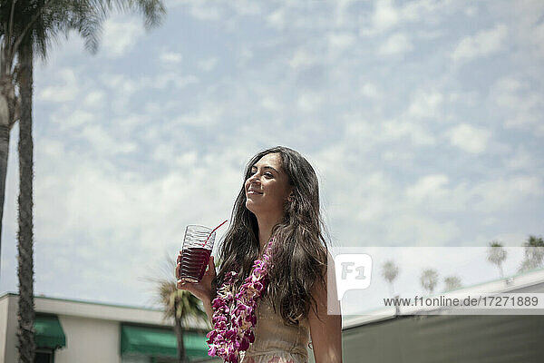 Junge Frau steht mit Saftgetränk und trägt eine Blumengirlande vor bewölktem Himmel