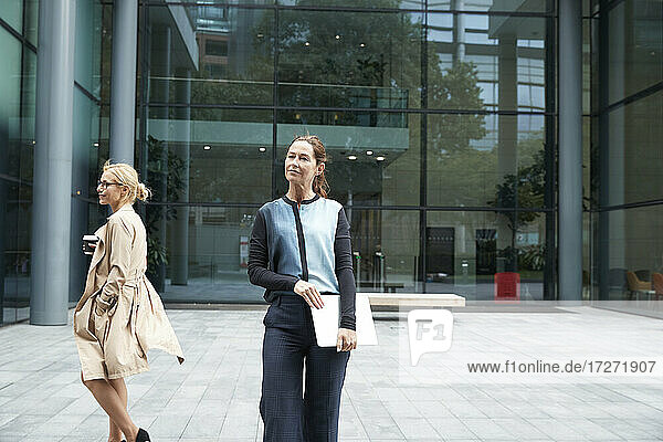 Woman standing and holding laptop with colleague walking in background against office building