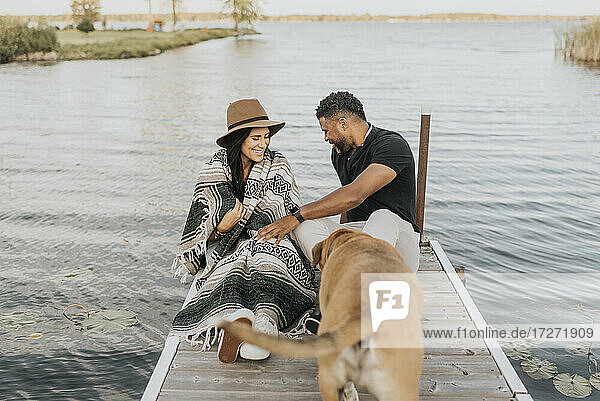 Lächelnde Frau in Decke sitzt bei Mann und Hund auf einem Steg am See