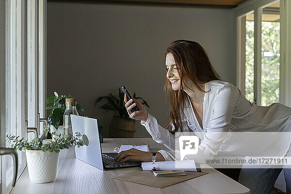 Smiling psychologist using mobile phone while working on laptop at home