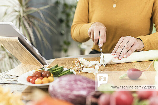 Frau schneidet Gemüse  während sie mit einem digitalen Tablet auf dem Tisch zu Hause steht