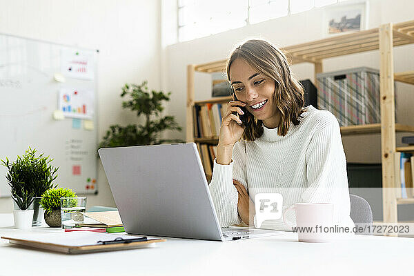 Smiling businesswoman talking on mobile phone while working on laptop at office