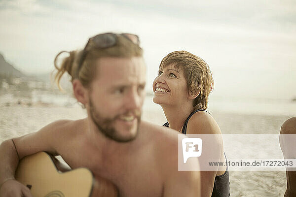 Lächelnder Mann spielt Gitarre am Strand sitzend an einem sonnigen Tag