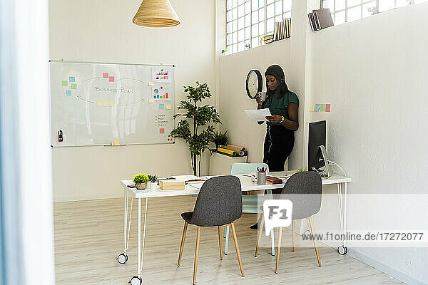 Geschäftsfrau mit Kaffeetasse beim Lesen von Papier  während sie sich im Büro an die Wand lehnt