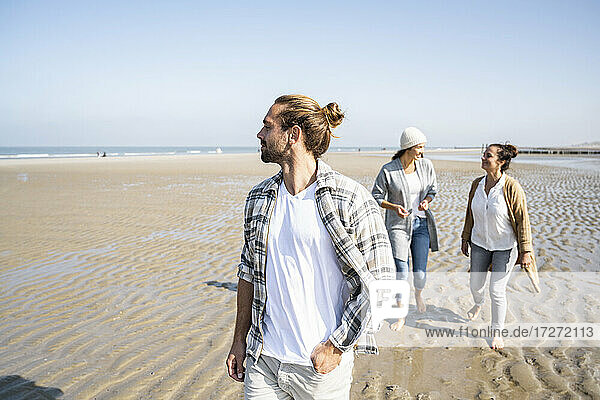 Mann schaut weg  während er mit Frauen im Hintergrund am Strand spazieren geht