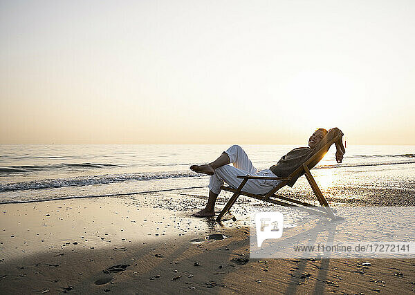 Lächelnde junge Frau entspannt sich auf einem Klappstuhl am Strand gegen den klaren Himmel bei Sonnenuntergang