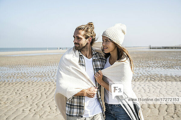 Nachdenkliches junges Paar  das in eine Decke gehüllt ist und wegschaut  während es am Strand an einem sonnigen Tag steht