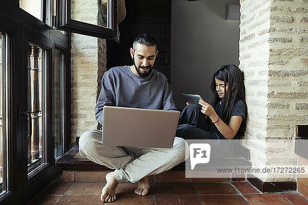 Girl using digital table while sitting by father working on laptop at home