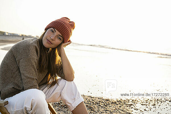Schöne junge Frau mit Strickmütze sitzt am Strand bei Sonnenuntergang