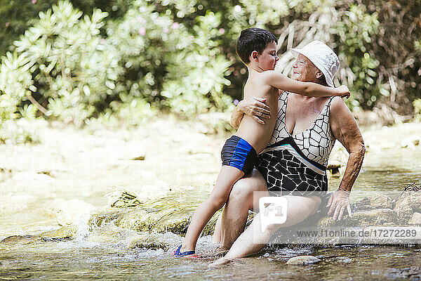 Senior woman looking at grandson standing with arm around her in river at forest
