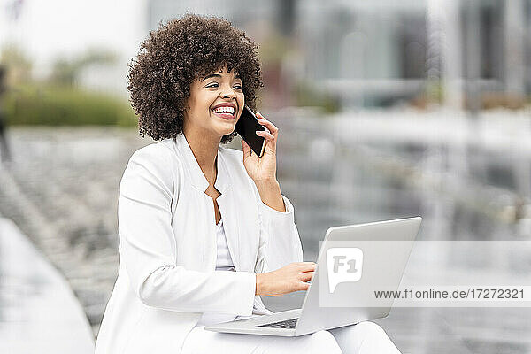 Lächelnde Geschäftsfrau  die auf einer Bank im Freien sitzt und mit ihrem Smartphone spricht