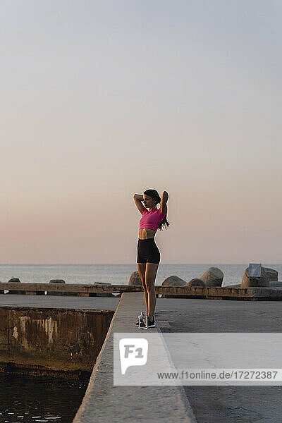 Junge Frau mit Hand in den Haaren steht auf einer Stützmauer am Meer