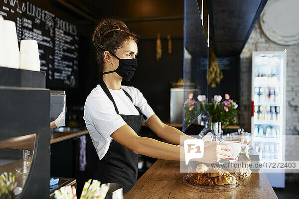 Weibliche Barista mit Gesichtsschutzmaske  die Kaffee ausschenkt  während sie in einem Café arbeitet