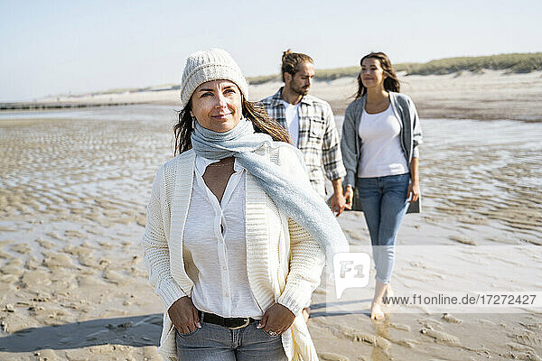 Smiling woman looking away while walking with couple holding hands in background at beach
