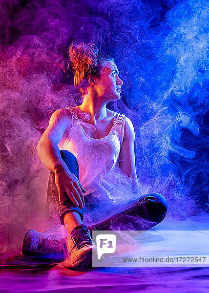 Junge Frau schaut weg  während sie inmitten von rosa und blauem Rauch im Studio sitzt