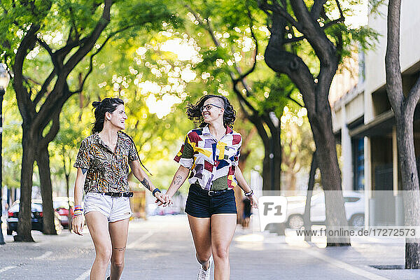Happy lesbian couple holding hands and running on footpath in city during sunny day