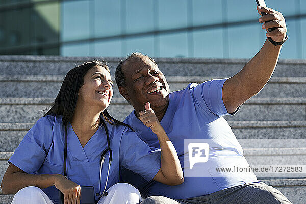 Lächelnde Ärzte  die ein Selfie machen  während sie auf der Treppe vor dem Krankenhaus sitzen