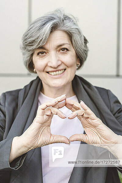 Smiling businesswoman making heart with finger while standing against wall