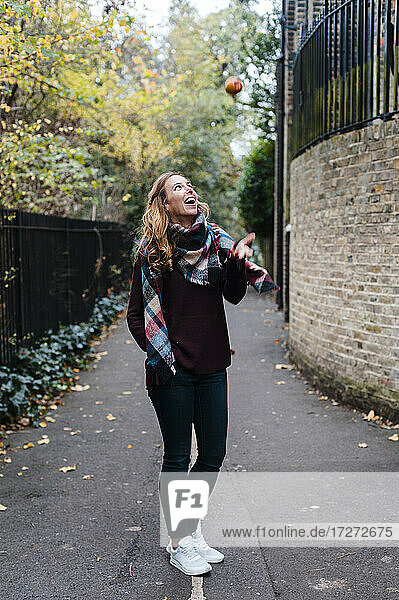 Smiling woman catching apple while standing on footpath during autumn