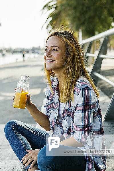 Junge Frau  die wegschaut  während sie auf einem Fußweg in der Stadt sitzt und Saft trinkt