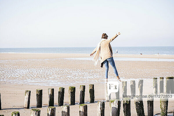 Junge Frau  die es genießt  an einem sonnigen Tag am Strand auf Holzpfählen mit ausgestreckten Armen zu laufen