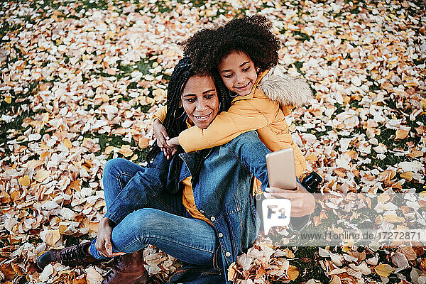 Mädchen  das seine Mutter umarmt und ein Selfie mit dem Smartphone macht  während es auf Herbstblättern sitzt