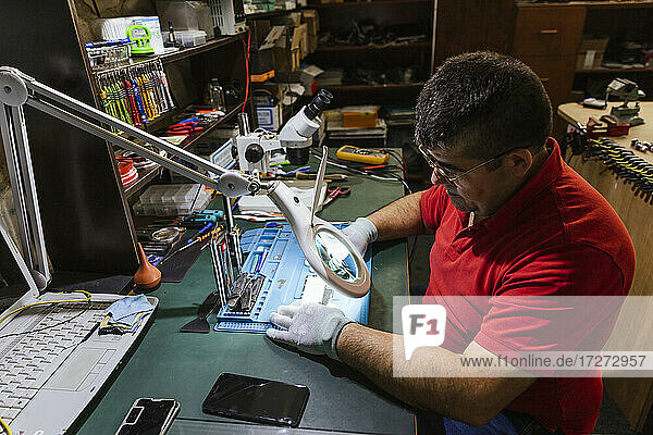 Arbeiter  der ein kaputtes Smartphone durch ein Vergrößerungsglas in einer Reparaturwerkstatt betrachtet
