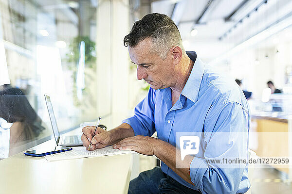 Selbstbewusster männlicher Berufstätiger  der mit seinem Laptop in einem Café sitzt und auf Papier schreibt
