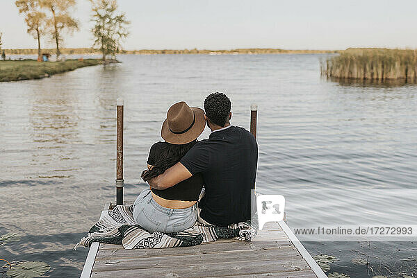 Couple with arm around admiring view while sitting on pier against lake