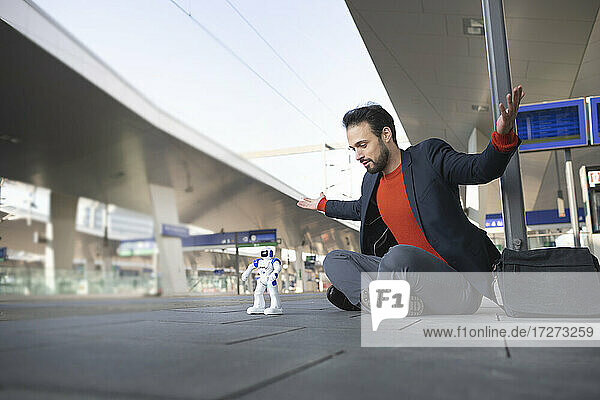Confident entrepreneur with arms outstretched looking at robot while sitting on railroad platform