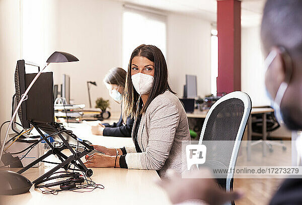 Weiblicher Angestellter mit Gesichtsmaske  der einen Kollegen ansieht  während er am Schreibtisch im Büro sitzt  während COVID-19