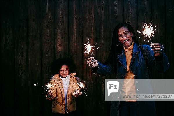 Smiling mother and daughter playing with sparkler while standing against wall