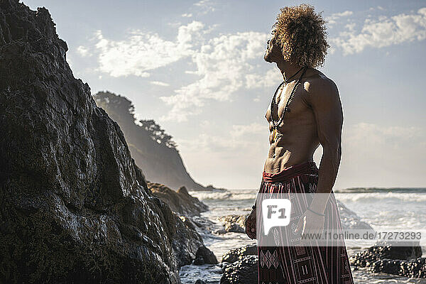 Shirtless muscular man standing by rock formations at beach against sky