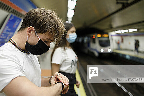 Junger Mann  der die Zeit prüft  während er neben einer Frau mit sozialer Distanz am U-Bahn-Bahnsteig steht
