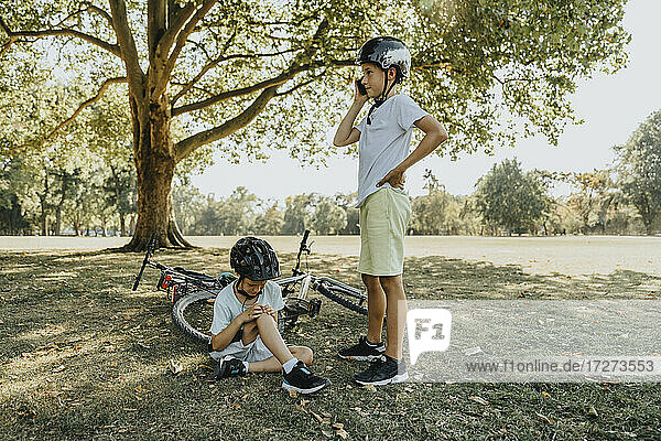Boy talking on smart phone while brother holding hurt sitting in public park
