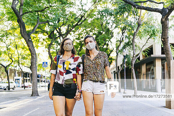 Lesbisches Paar mit Gesichtsschutzmaske hält sich auf dem Fußweg an den Händen