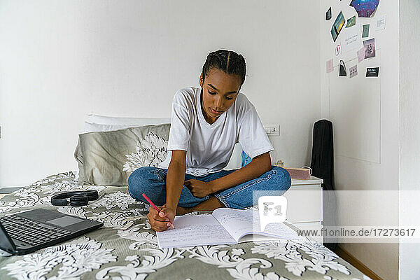 Concentrating teenage girl girl writing in exercise book while sitting on bed in bedroom at home