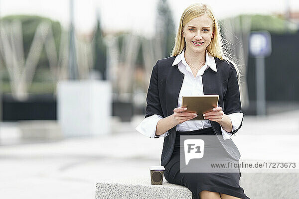 Young businesswoman using digital tablet while sitting on bench against building