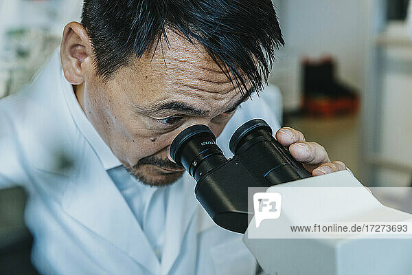 Male scientist looking though microscope while man standing in background at laboratory
