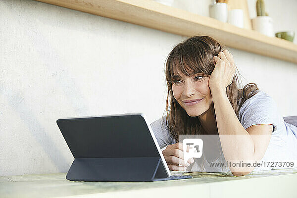 Lächelnde reife Frau mit Händen in den Haaren  die ein digitales Tablet benutzt  während sie zu Hause auf der Vorderseite liegt