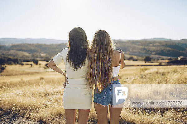Weibliche Freunde betrachten die Aussicht  während sie auf einem grasbewachsenen Feld gegen den Himmel an einem sonnigen Tag stehen