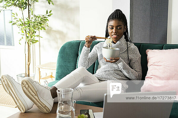 Junge Frau isst Nudeln  während sie einen Film auf dem Laptop im Wohnzimmer zu Hause ansieht