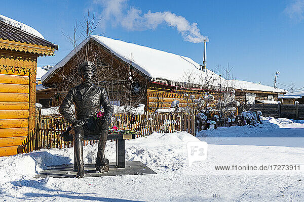 Russland  Republik Sacha  Jakutsk  Statue eines auf einer Bank sitzenden Soldaten vor rustikalen Häusern im Winter