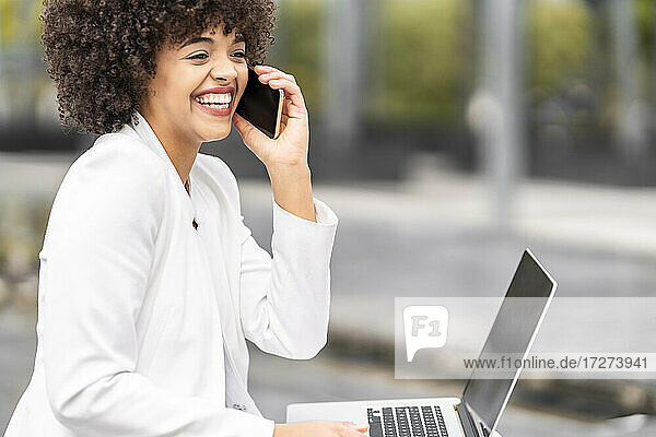 Lächelnde Geschäftsfrau  die auf einer Bank im Freien sitzt und mit ihrem Handy telefoniert