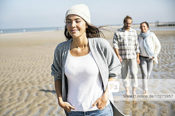 Lächelnde Frau  die mit ihrem Freund und ihrer Mutter im Hintergrund am Strand spazieren geht