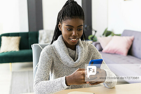 Smiling young woman text messaging on smart phone while sitting in living room at home