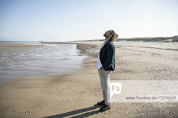 Junger Mann mit Händen in den Taschen am Strand stehend gegen den klaren Himmel