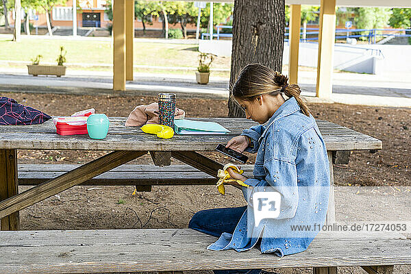 Teenager-Mädchen  das ein Bild von einer Banane macht  während es auf einer Bank in einem öffentlichen Park an einem sonnigen Tag sitzt
